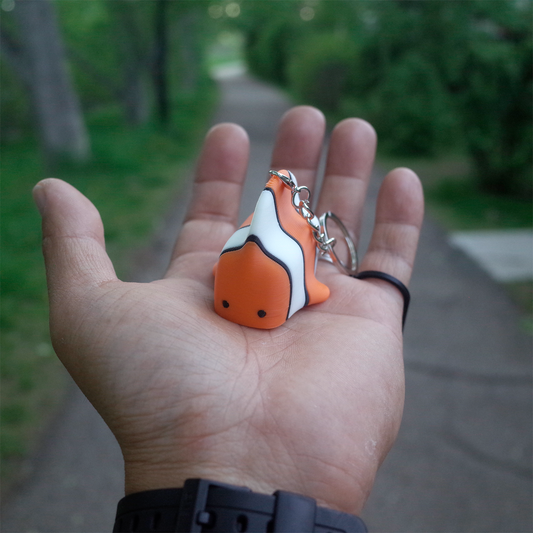A hand holding a cute orange and white 3D printed clownfish keychain with a silver chain, facing front with a soft expression.