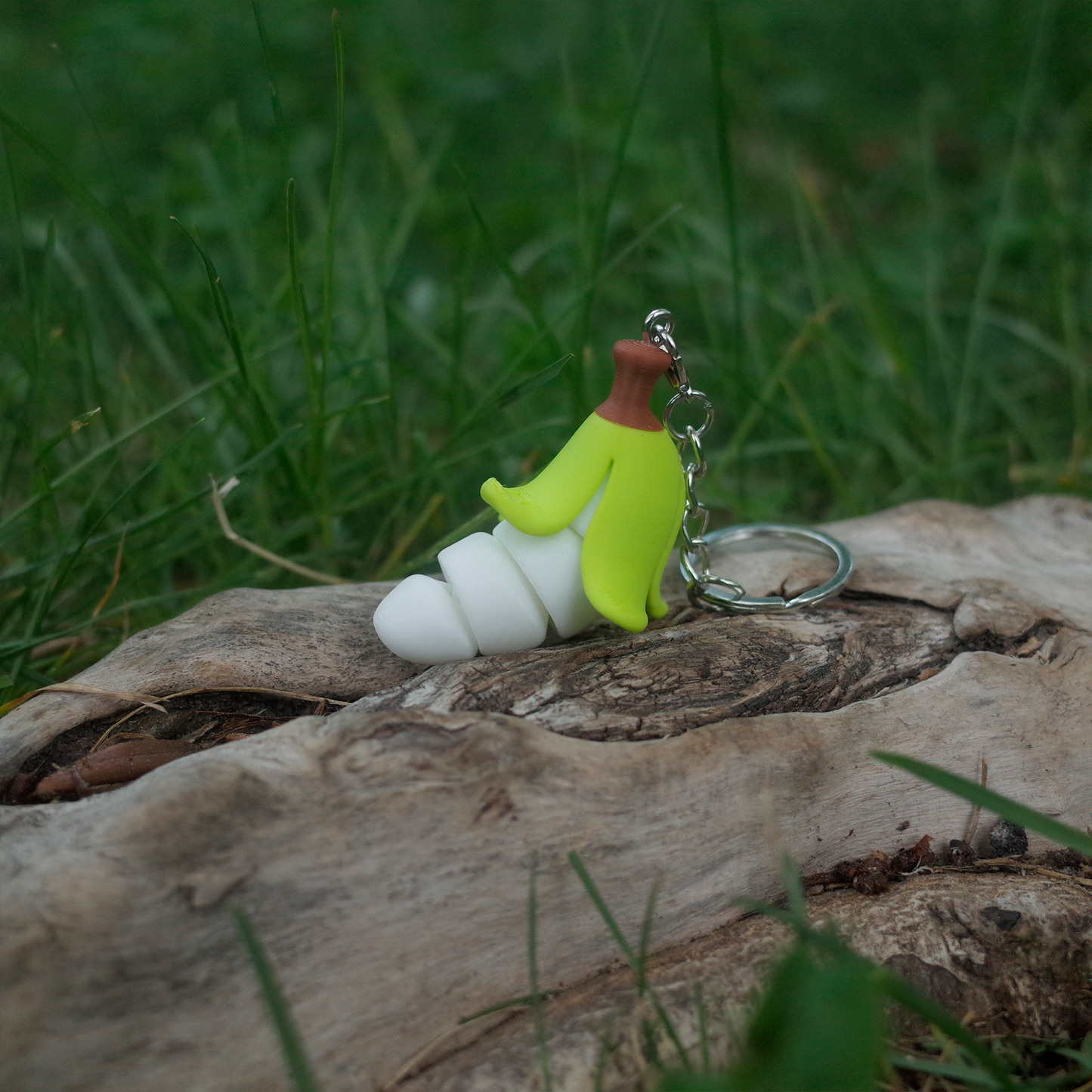 A 3D printed chubby banana keychain with a light green peel and white curvy body, resting on wood in the grass.