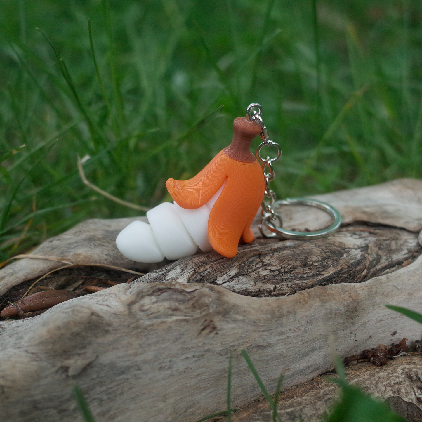 A vivid orange peeled chubby banana charm keychain resting on a log with green grass in the background.