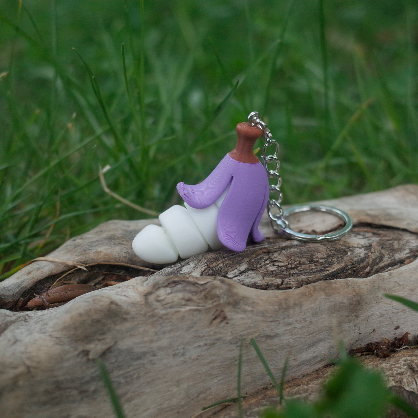 Cute 3D printed keychain shaped like a banana with a purple peel and chubby white body, placed on a driftwood log.