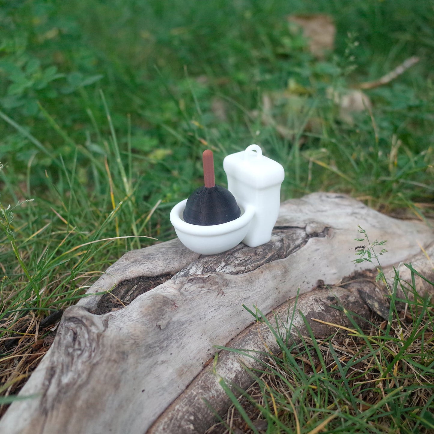 Miniature white toilet toy with a black plunger sitting on a log outdoors