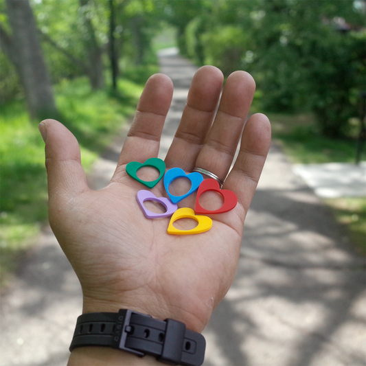 A hand holding five vibrant heart-shaped rings in red, yellow, green, blue, and purple, with a tree-lined path in the background.