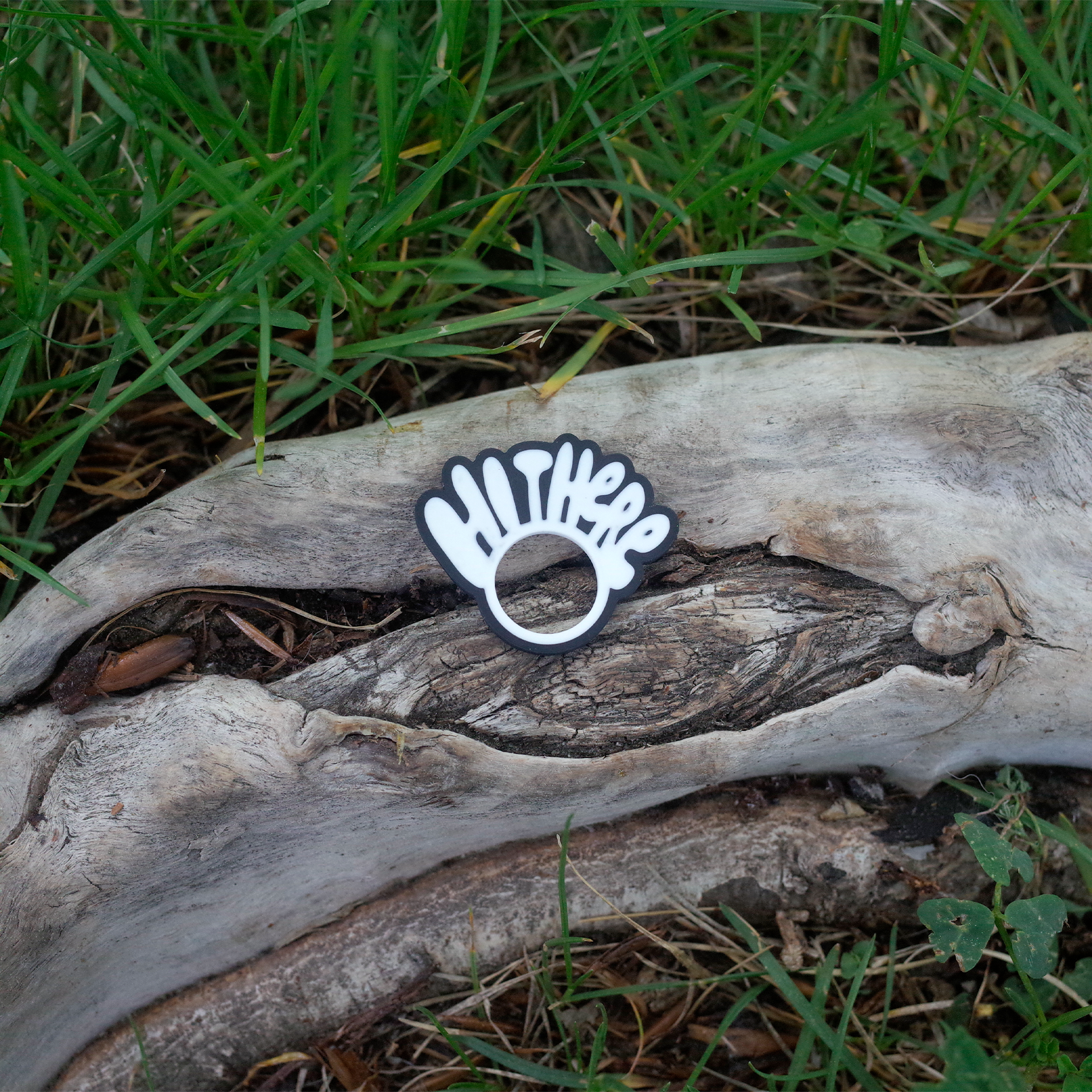 A black and white 3D printed charm shaped like a waving hand with the words “Hi There”, placed on a textured tree trunk surrounded by grass.