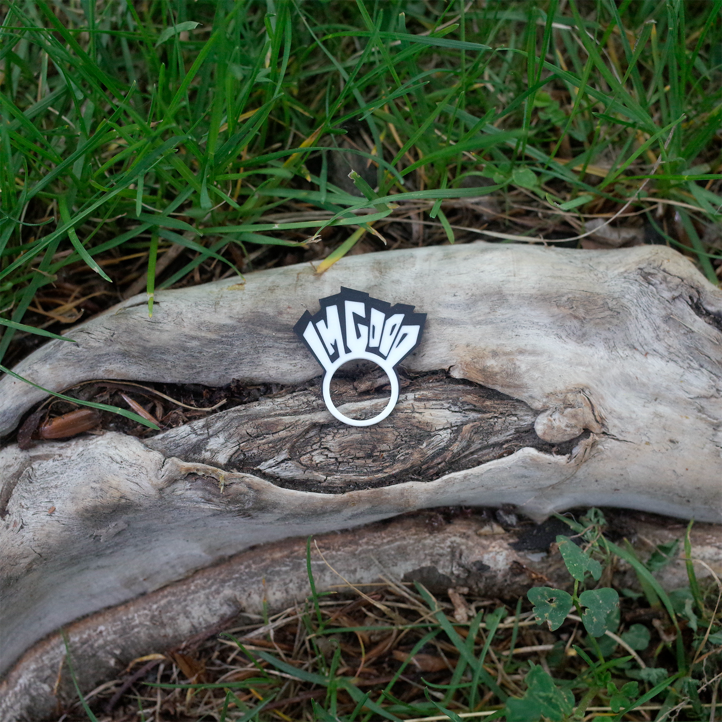 A black and white 3D printed charm that reads “I’M GOOD”, shaped like a tiny sign with bold blocky letters, placed on a weathered tree trunk surrounded by grass.