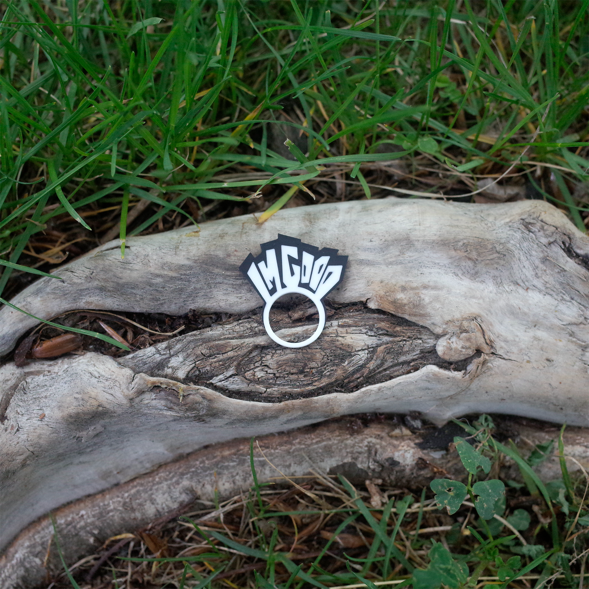 A black and white 3D printed charm that reads “I’M GOOD”, shaped like a tiny sign with bold blocky letters, placed on a weathered tree trunk surrounded by grass.