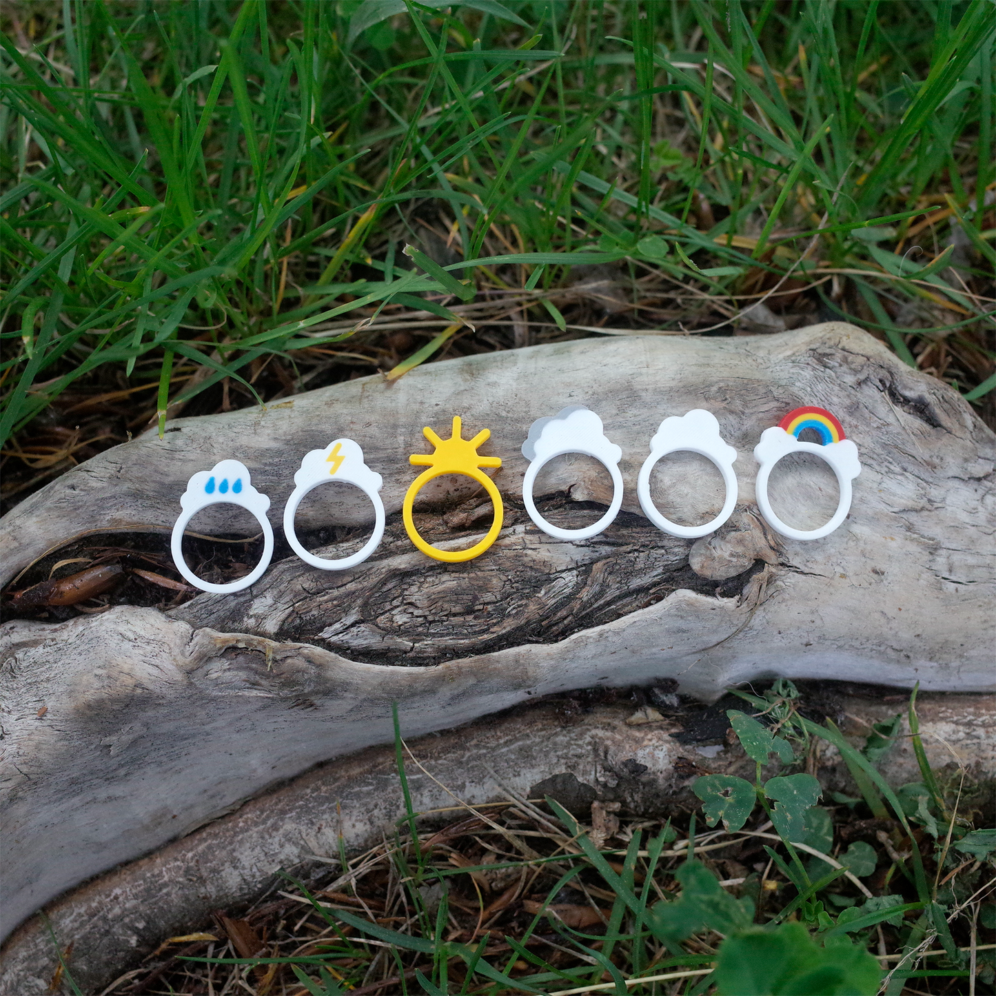 Six weather-themed 3D printed rings arranged on a tree trunk, featuring sun, clouds, rain, lightning, and rainbow designs.