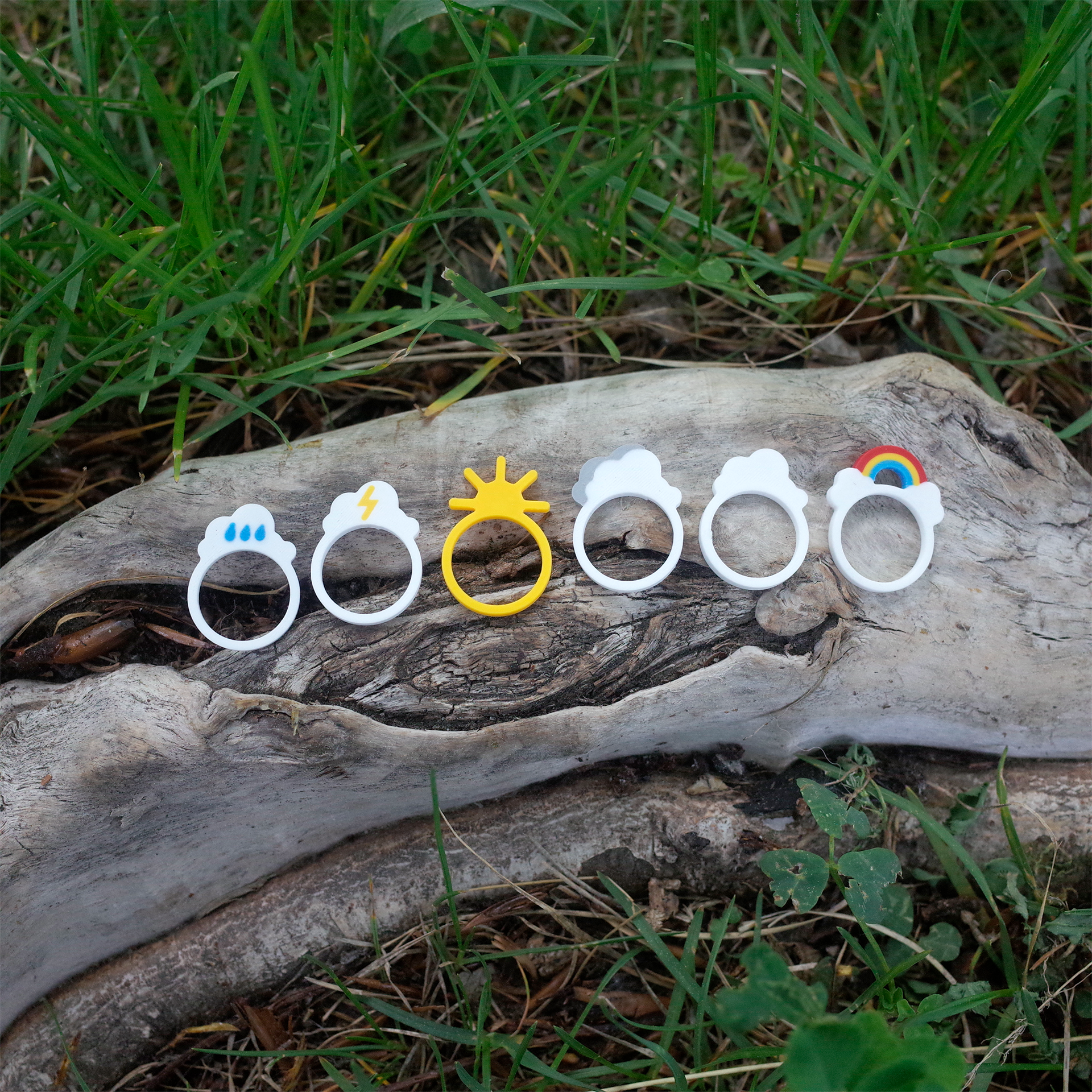 Six weather-themed 3D printed rings arranged on a tree trunk, featuring sun, clouds, rain, lightning, and rainbow designs.