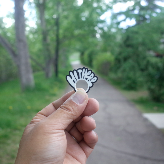 A hand holding a small black and white charm shaped like a waving “Hi There” sign, with a green park path in the background.