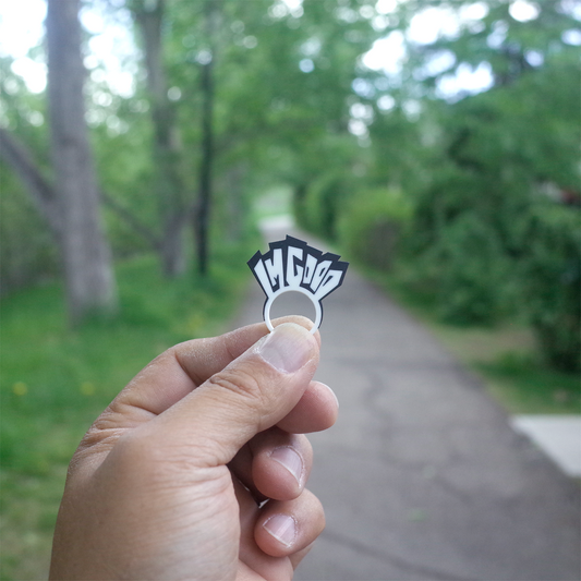 A hand holding a small “I’M GOOD” 3D printed charm in front of a tree-lined path, with soft green background and natural light.