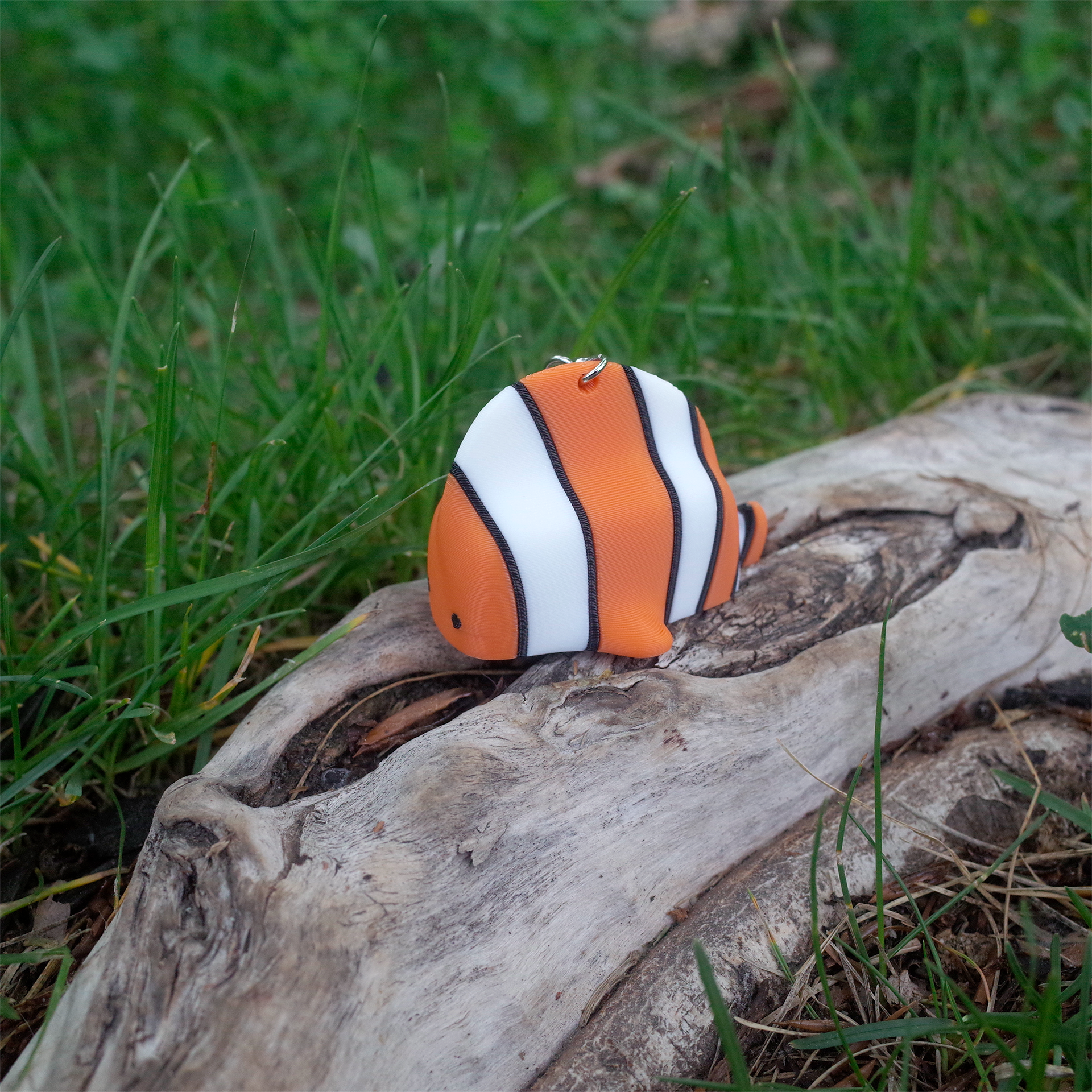 A 3D printed orange and white clownfish charm with black stripes, resting on a piece of driftwood surrounded by grass.