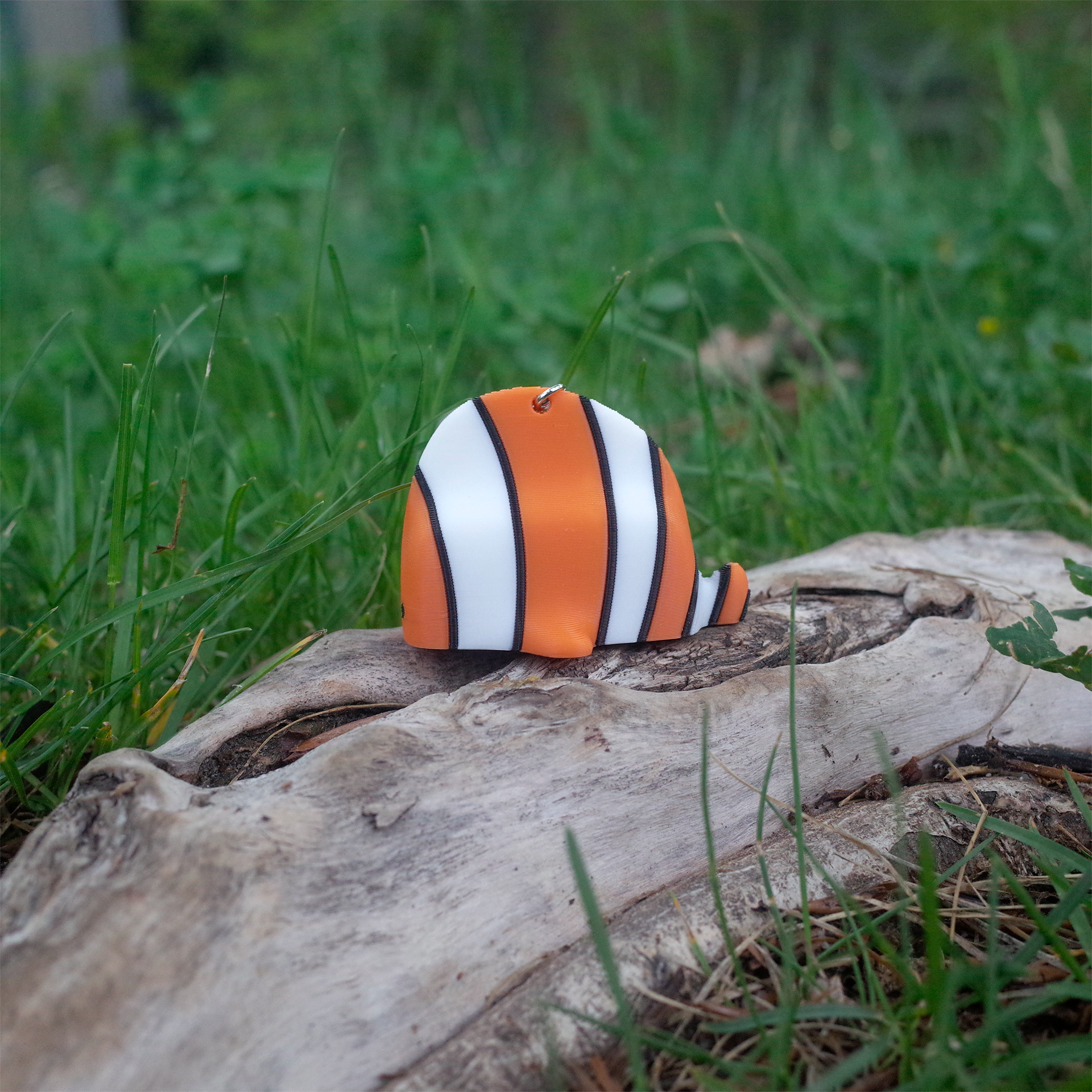 Side view of a plump clownfish-shaped 3D printed charm, featuring orange, white, and black stripes, set on outdoor wood.
