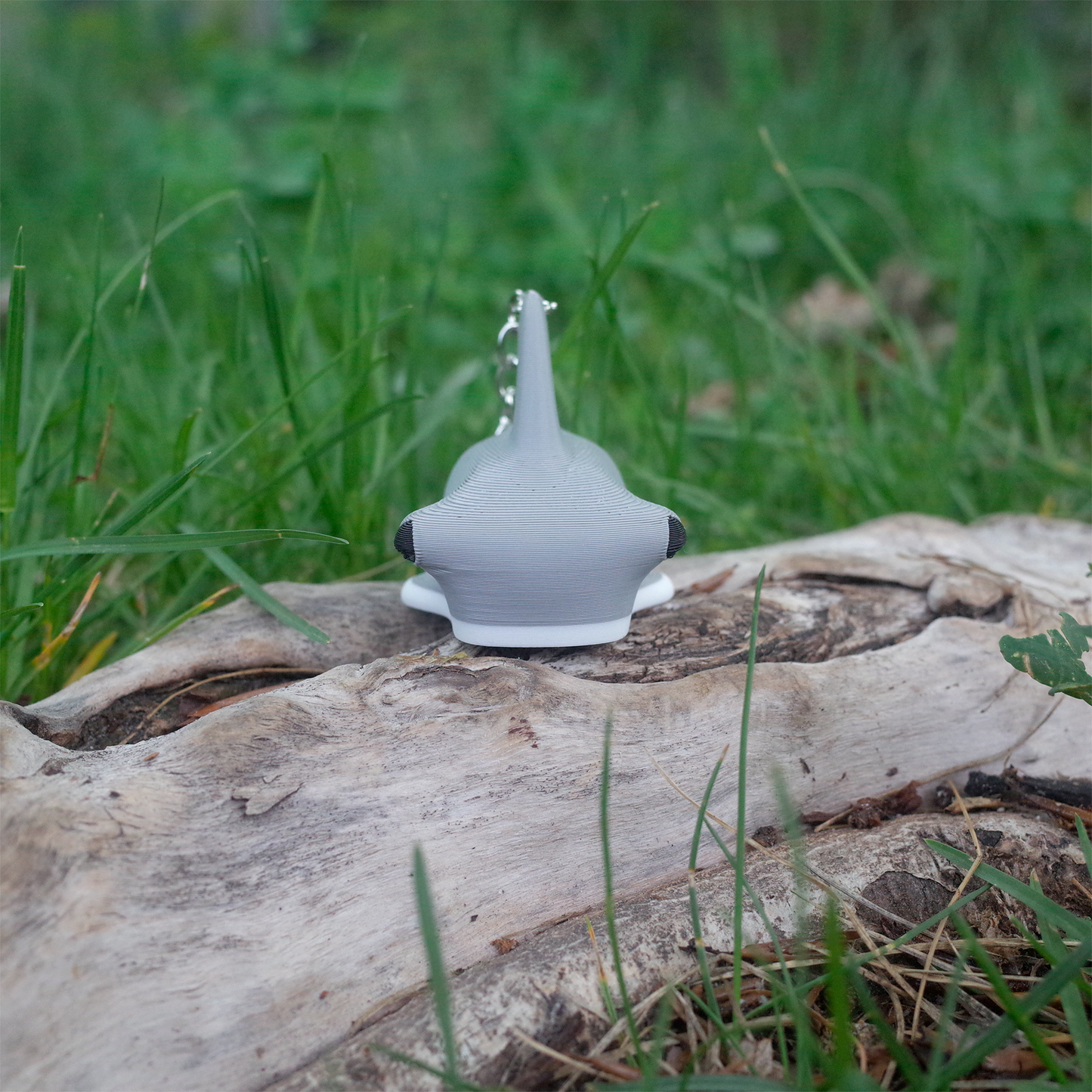 A front-facing 3D printed hammerhead shark keychain, gray with white underside, sitting upright on a tree log in the grass.