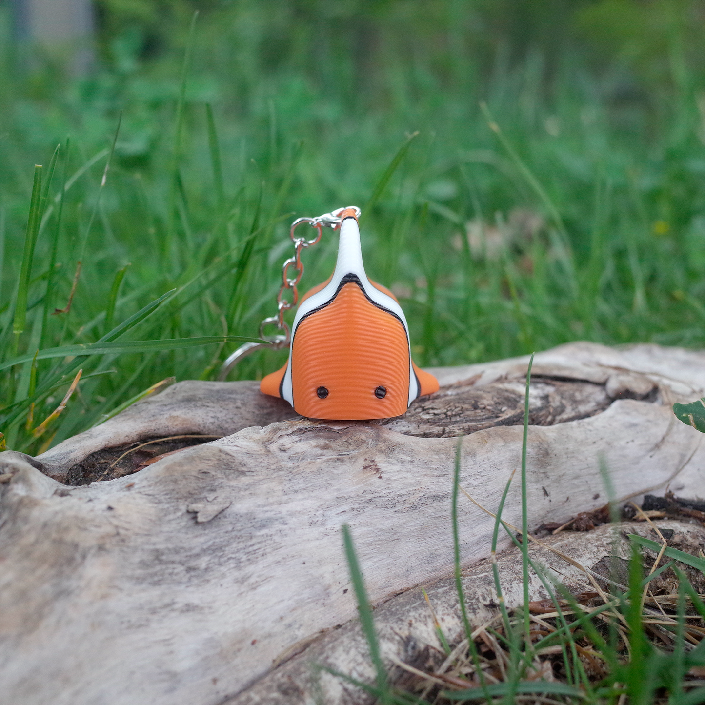 Front view of the 3D printed clownfish charm, with its tiny dot eyes and rounded body, standing upright on a tree log.