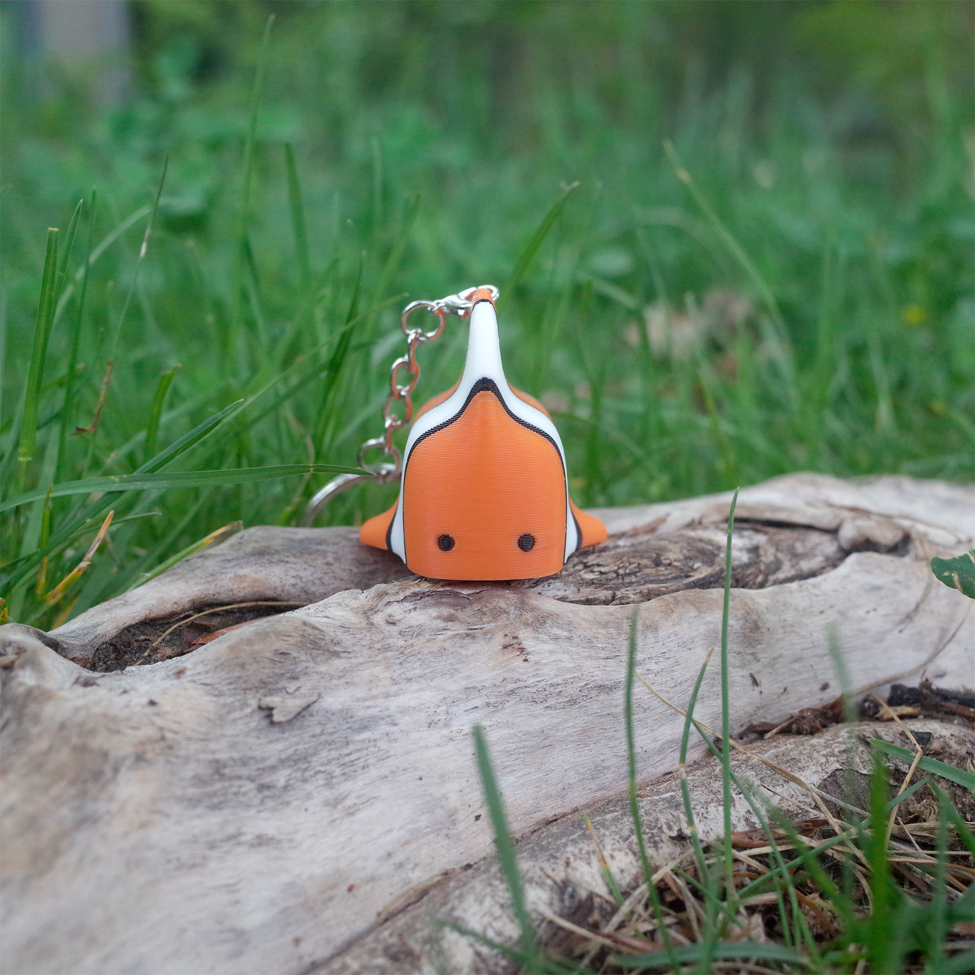 Front view of the 3D printed clownfish charm, with its tiny dot eyes and rounded body, standing upright on a tree log.