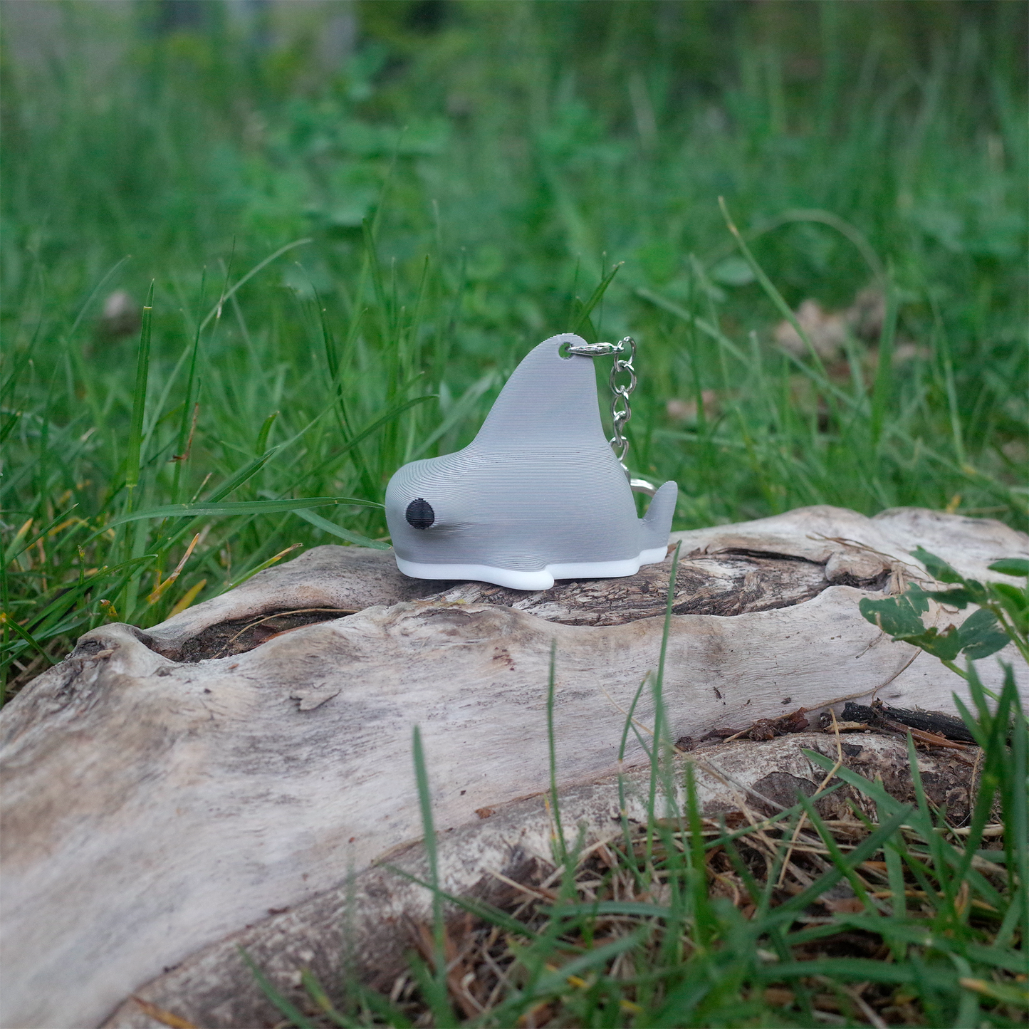 Side view of the hammerhead shark charm with a silver keychain ring, resting on a tree trunk with green grass background.