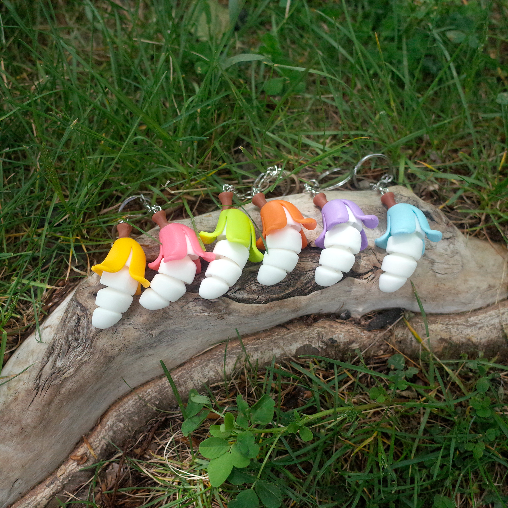 A full lineup of rainbow Chubby Bananaz charms resting on a log in the grass, each banana sporting a unique peel.