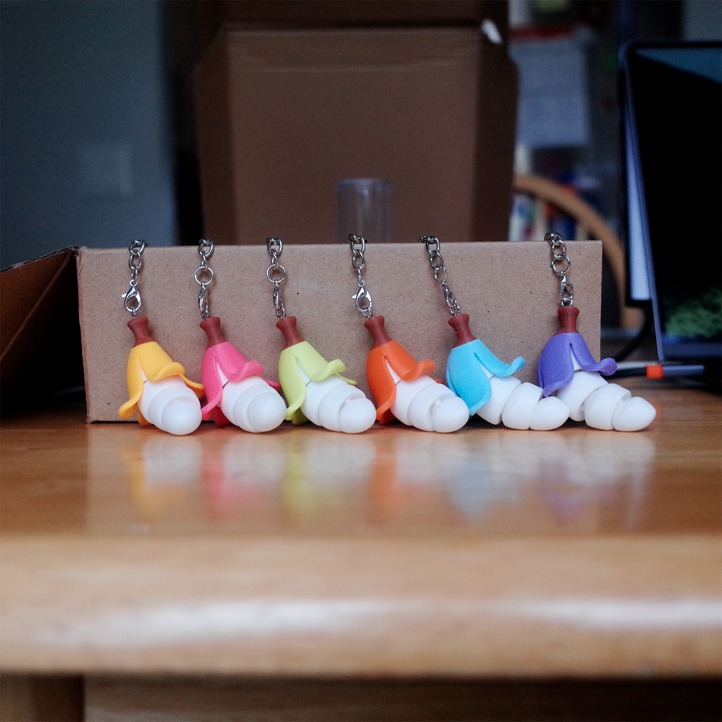 Seven colorful Chubby Bananaz keychains lined up in a row on a cardboard box, each with a different peel color.