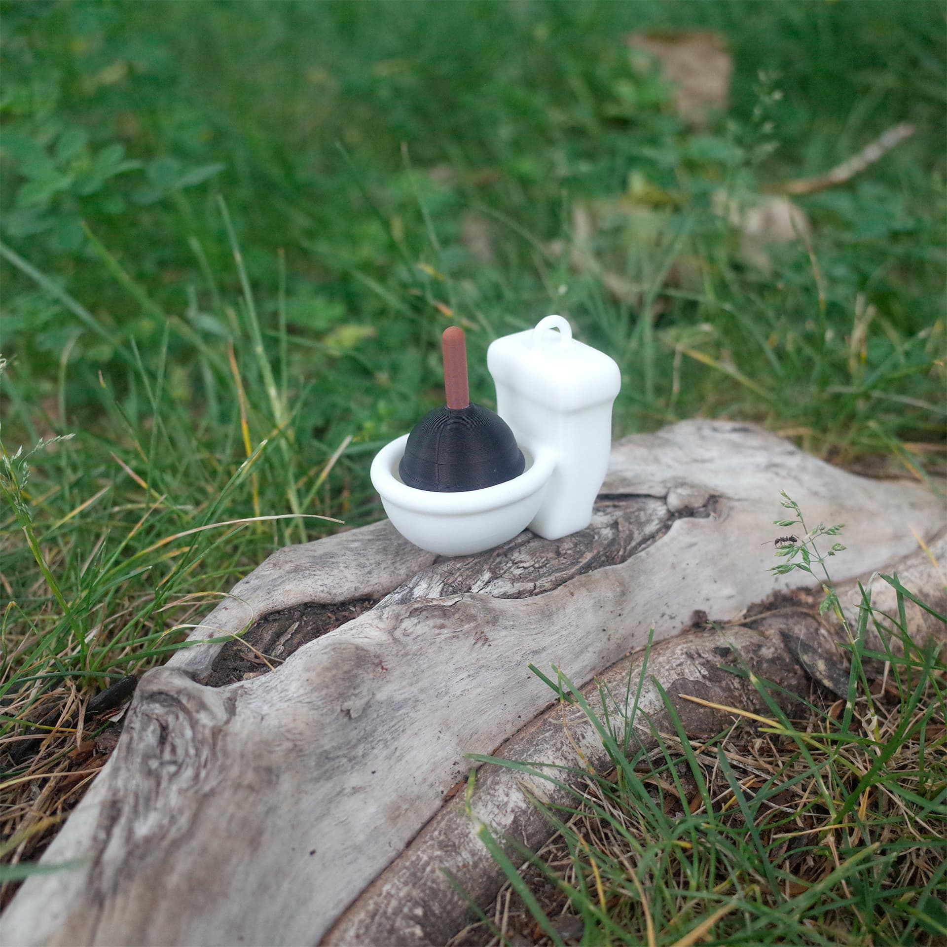Miniature white toilet toy with a black plunger sitting on a log outdoors