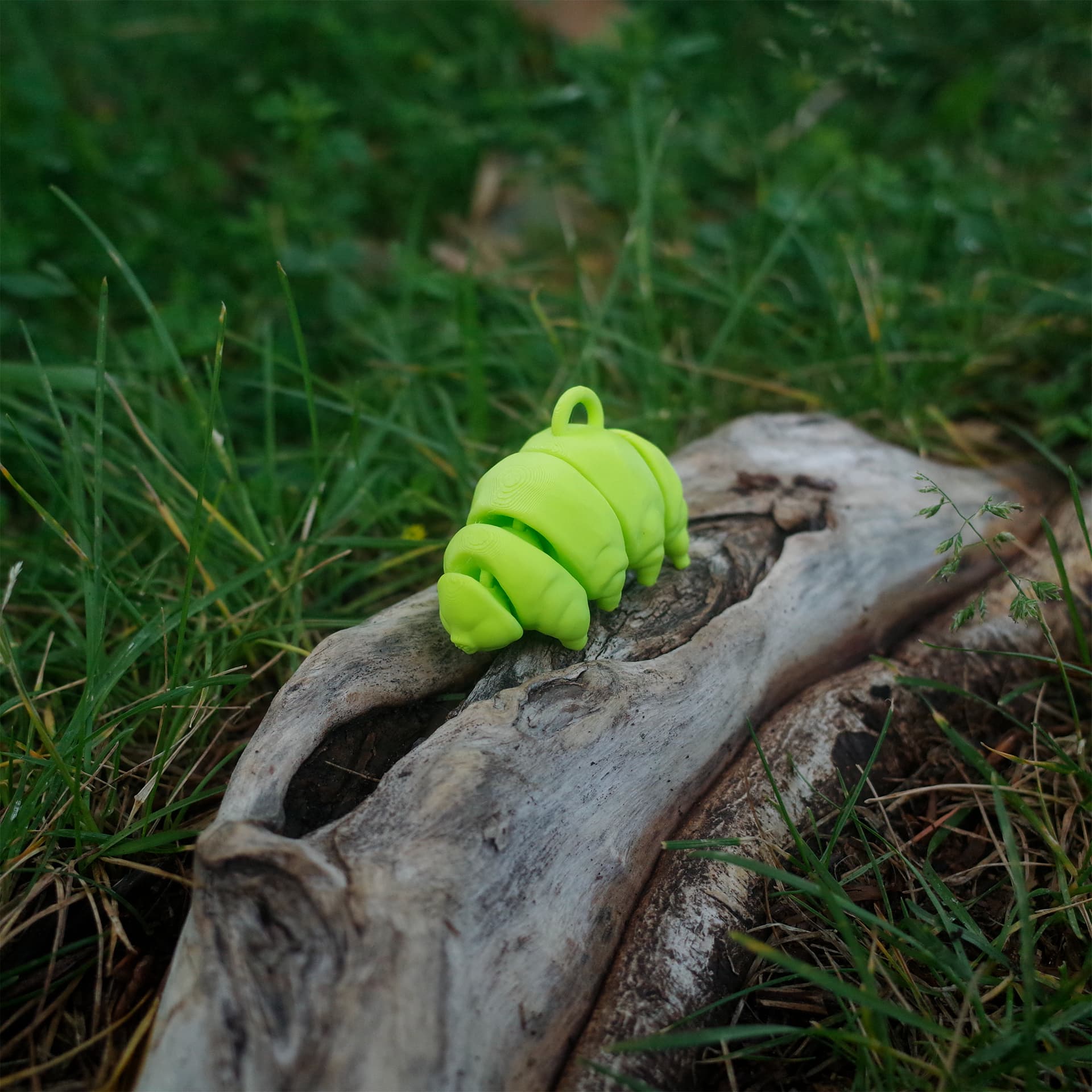 A bright green 3D printed tardigrade figurine resting on a piece of driftwood in a grassy outdoor setting.