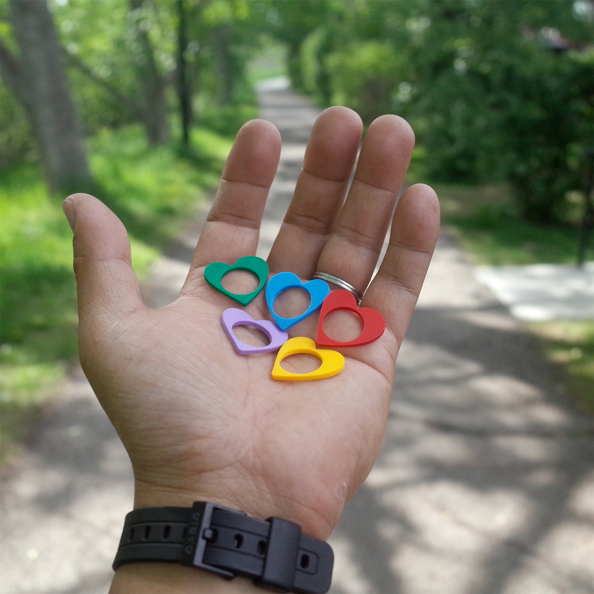 A hand holding five vibrant heart-shaped rings in red, yellow, green, blue, and purple, with a tree-lined path in the background.