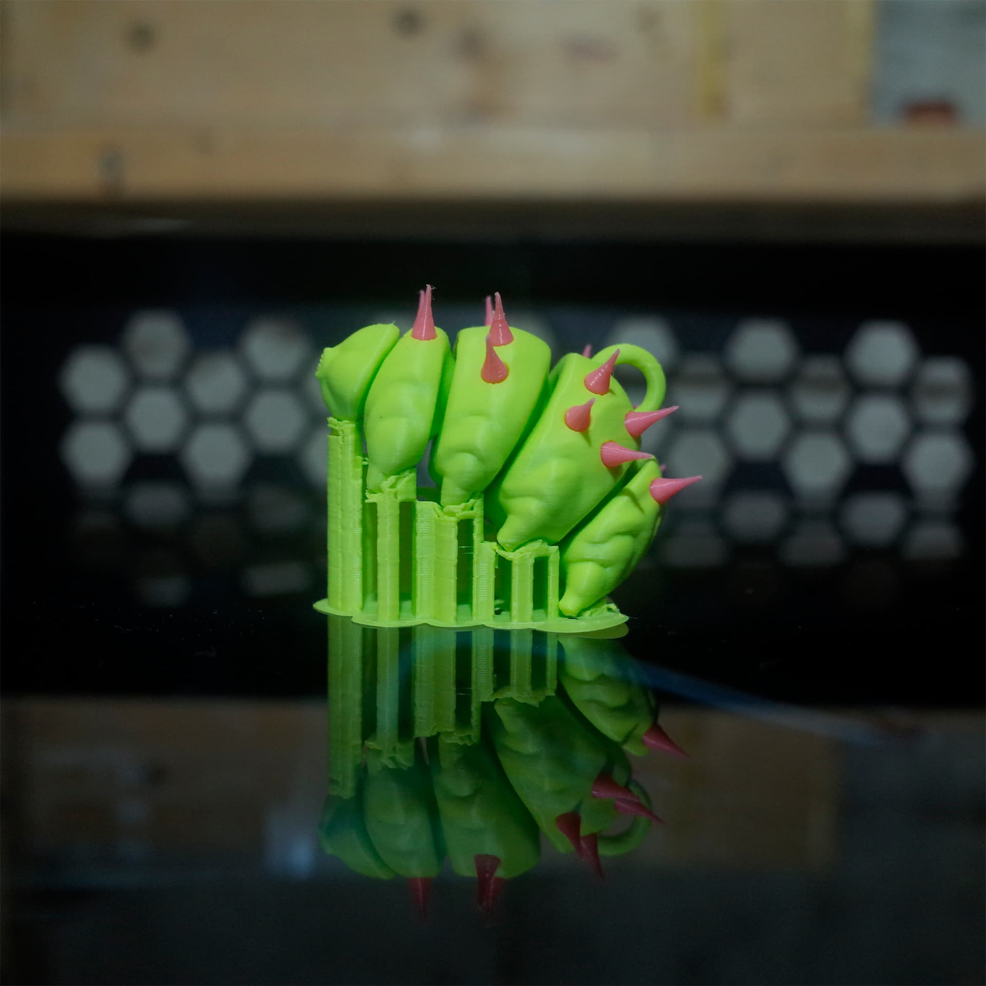 A punk-style green tardigrade toy with pink spikes, on a mirrored printer base.