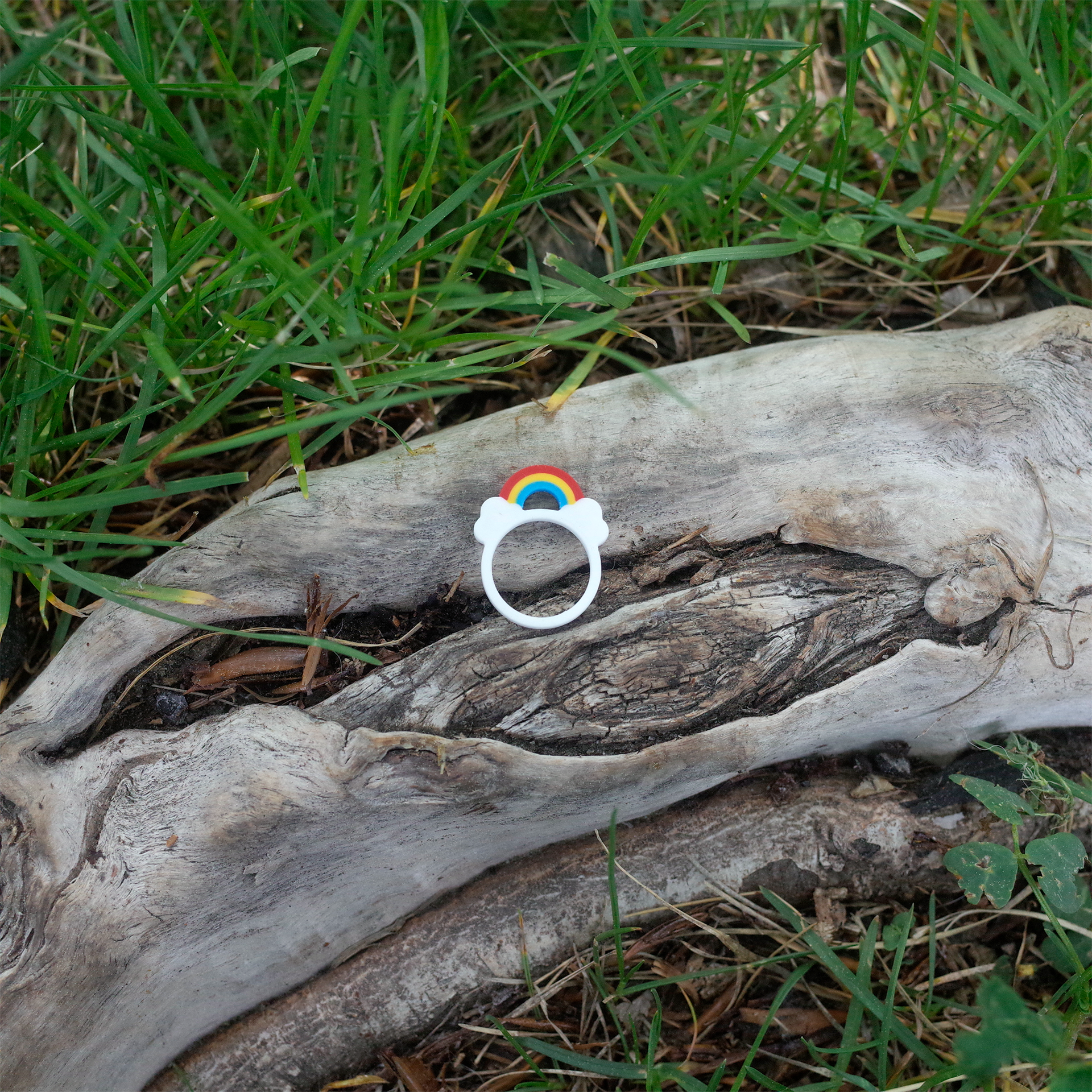 A white ring with a small rainbow over clouds, 3D printed and placed on a tree trunk among grass.