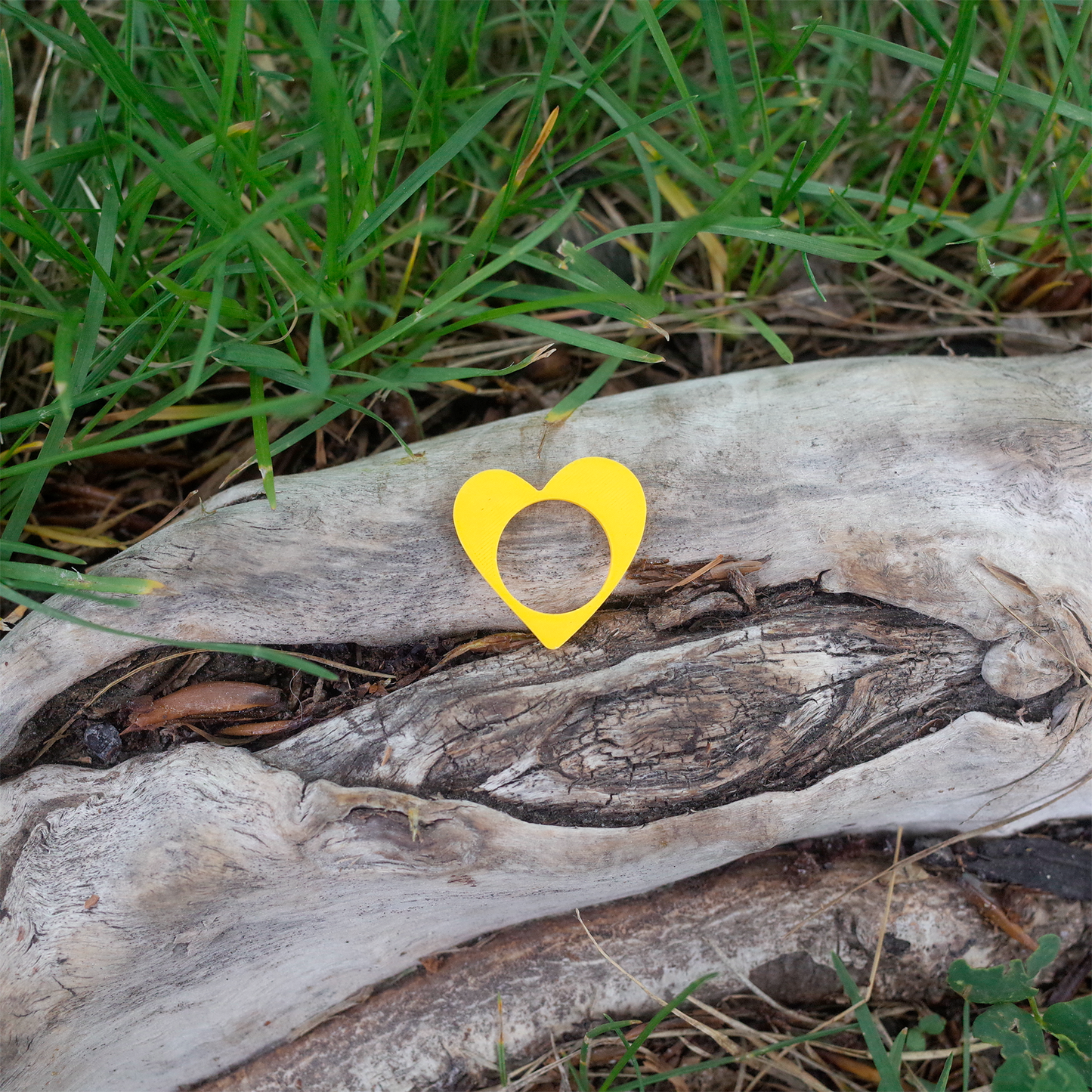 A bright yellow heart-shaped 3D printed ring placed on natural wood with grass surrounding.